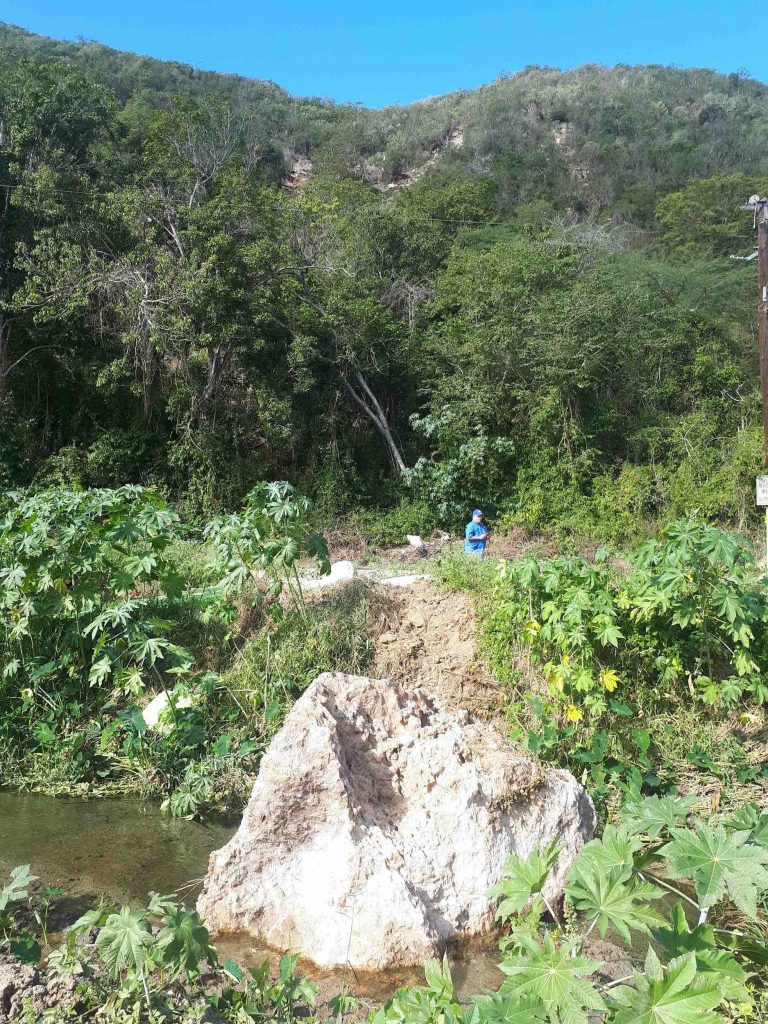 Volkswagen-sized boulder sourced from the upper part of the photo that landed in a small channel along road PR-335R in Barrio Barina of Yauco. Note multiple headscarps above, and the track of the boulder on the channel bank. Credit: Stephen Hughes